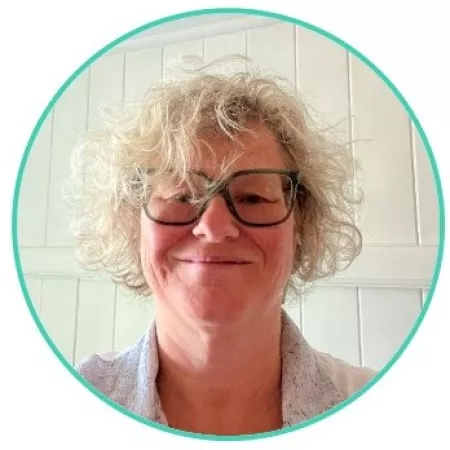 Head-and-shoulders portrait of a person with short curly light-coloured hair and dark-framed glasses, photographed against a light-coloured tiled wall. The image is cropped within a circular border.